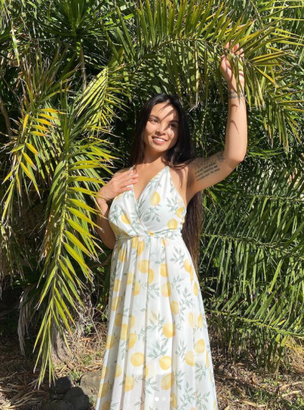 Girl holding up palm trees, smiling away from the camera. Wearing a lemon print maxi dress